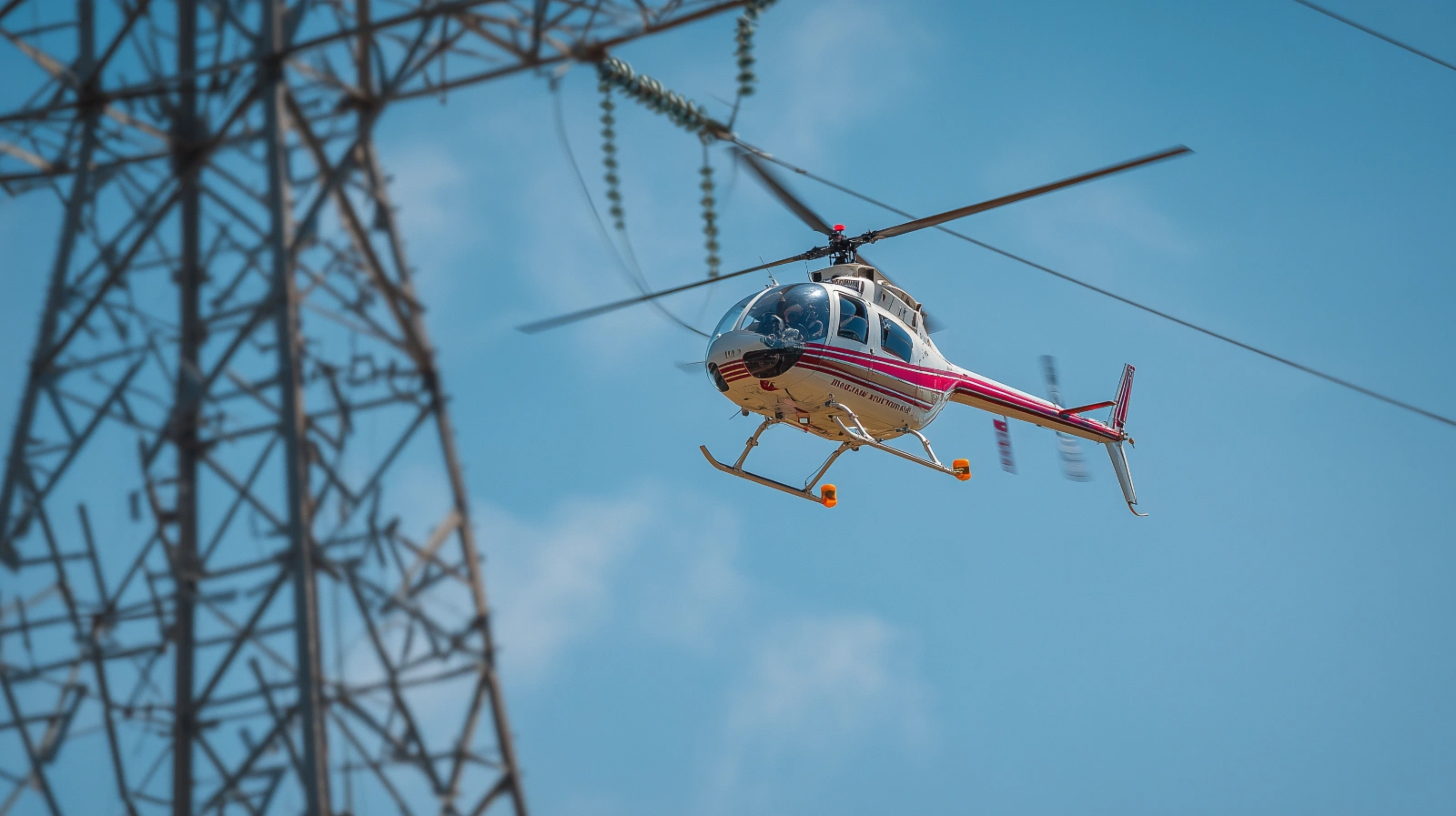 A Hughes 369D helicopter flying near a transmission tower and overhead utility lines in daylight.
